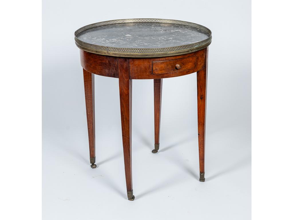 Table bouillote de style Louis XVI reposant sur un piètements gainé à roulette. Elle ouvre par deux tiroirs en ceinture et présente un plateau de marbre gris entouré d'une galerie ajourée en laiton. XIXème siècle. Hauteur : 75 cm. Diamètre : 66 cm.