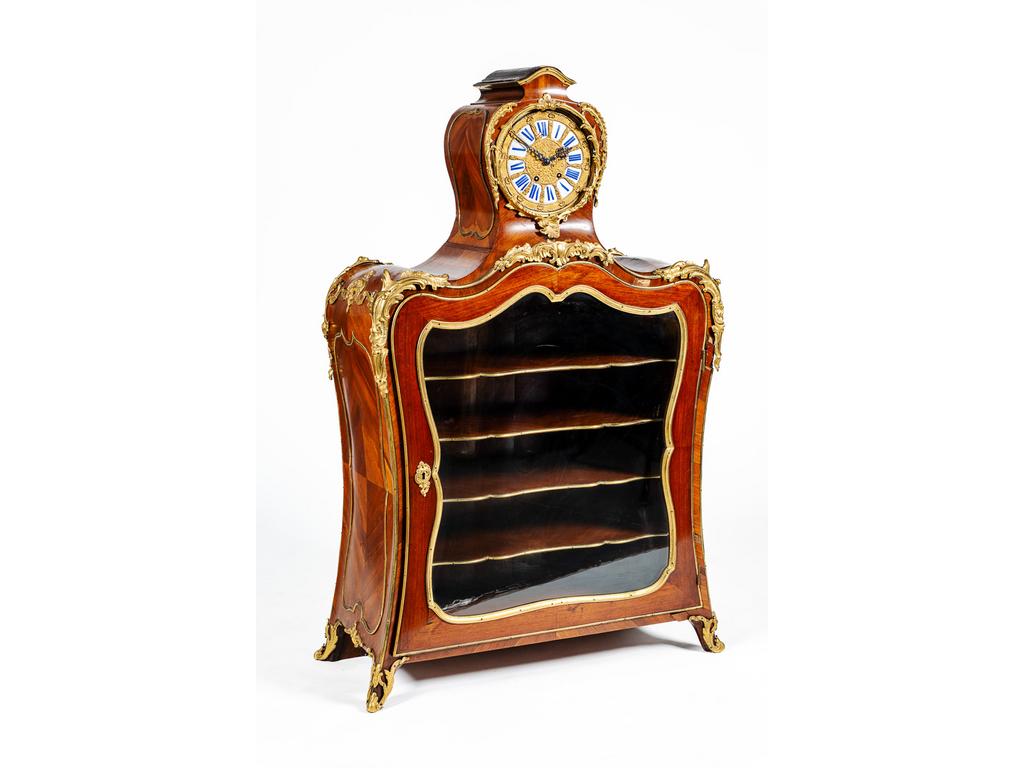 Petite Bibliothèque à Cartel dépoque Louis XV en bois de placage surmonté d'une pendule à chiffres romains. Belle ornementation de bronzes dorés à décor de feuilles d'acanthes, coquilles et agrafes. Elle ouvre en façade par une porte vitrée violonnée et repose sur deux petits pieds avant cambrés à sabots de bronze. (Manque la vitre au cadran, sauts au placage, gond à refixer). Dim : 139 x 85 x 38 cm.