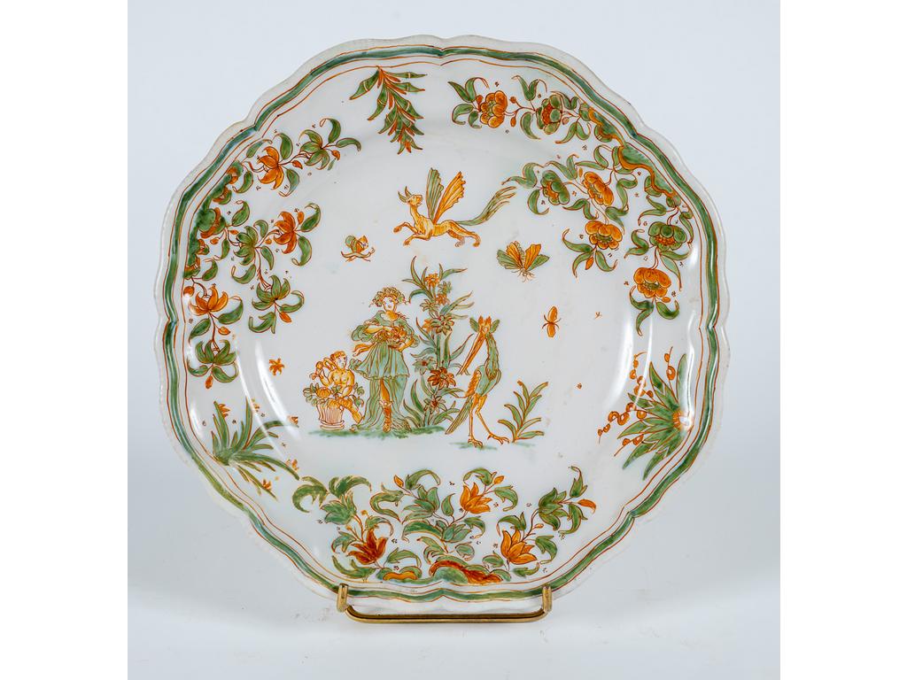 MOUSTIERS, Olérys & Laugier : Assiette en faïence polychrome à bords chantournés à décor de bacchantes et putti sur un tertre, dans un entourage feuillagé. Monogrammée au revers. Epoque XVIIIème siècle. (Restaurations).