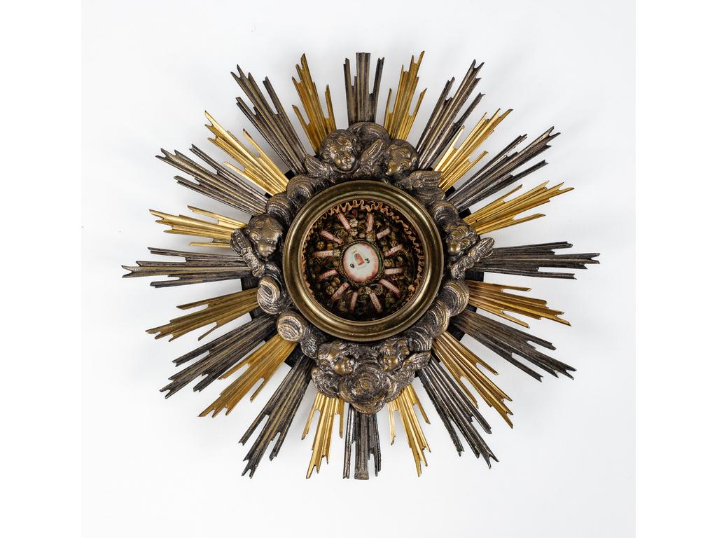 Petit reliquaire orné de paperolles dans un joli cadre en bronze et laiton argenté à décor rayonnant de tête d'angelots dans des nuées. Epoque XIXème siècle. Diamètre : 24.5 cm.