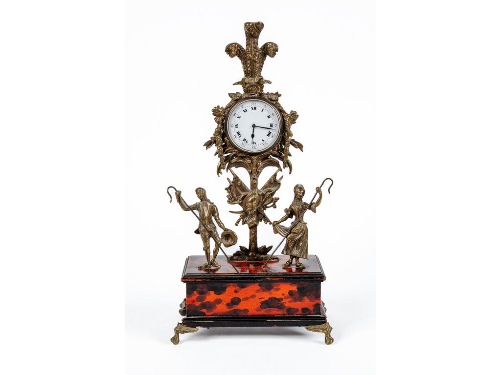 Porte montre en bronze et laiton à décor d'un couple de berger, trophée guirlandes de fleurs et panaches. Il repose sur un socle en bois peint à l'imitation de l'écaille, terminé par quatre petits pieds griffes de lion. Epoque fin XIXème siècle. (Eclats au cadran et manque une plainte au socle). Dim : 37 x 18.5 x 10 cm.