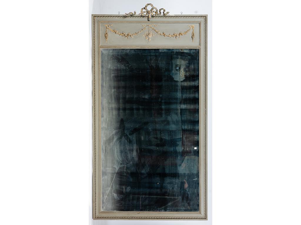 Grand miroir trumeau biseauté dans un encadrement de style Louis XVI en bois et stuc laqué gris et à décor de noeuds de rubans, guirlandes de fleurs et rehauts dorés. (Accidents et manques). Hauteur : 216 cm et Largeur : 115 cm.