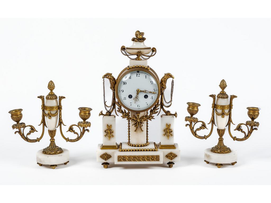 Garniture de cheminée de style Louis XVI en bronze et laiton et albâtre comprenant une pendule surmontée d'un vase fleuri, ainsi qu'une paire de flambeaux à deux bras de lumière. (Petits éclats, sans balancier et manque un élément de bronze sur chaque flambeaux). Dim de la pendule : 35 x 20.5 x 9 cm.