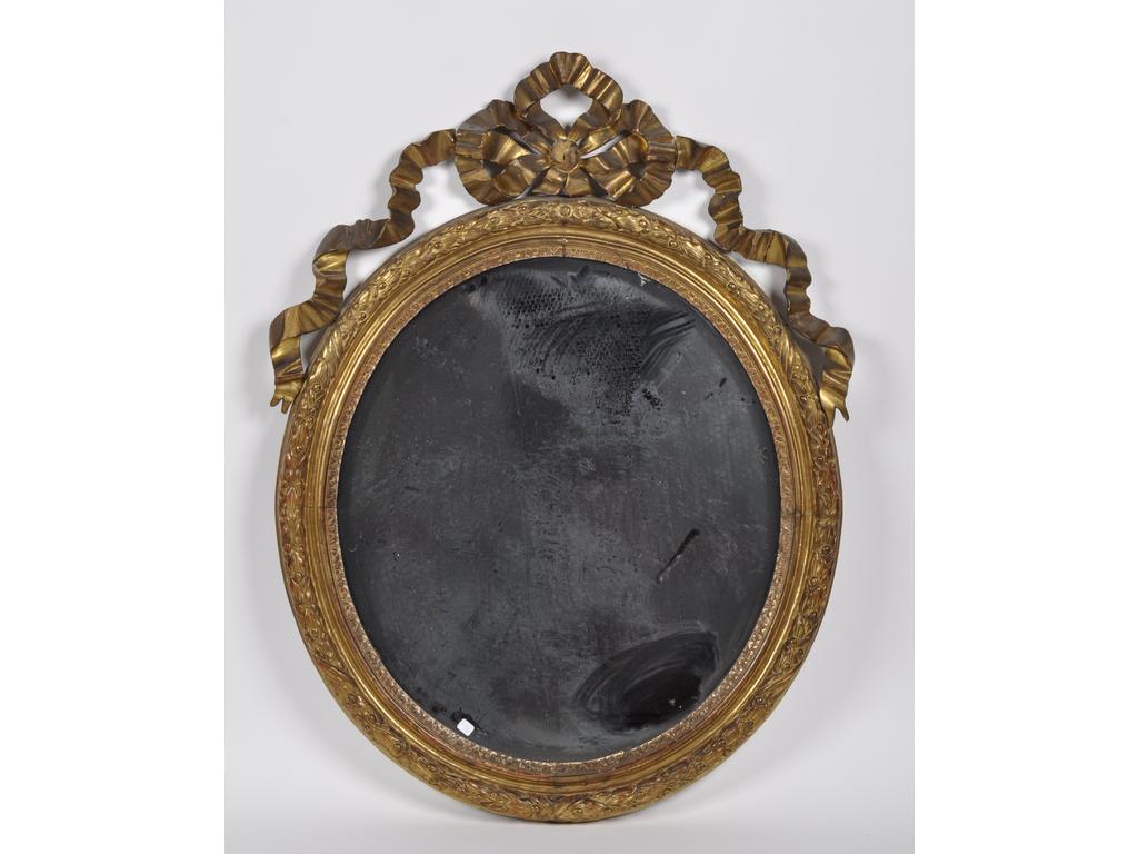 Miroir de forme ovale en bois et stuc à décor dans un encadrement à motif de feuilles de laurier, surmonté d'un grand noeud rubané. Epoque fin XIXème siècle. (Ruban abîmé, une partie cassée, éclats de dorure). Dim : 114 x 83 cm.