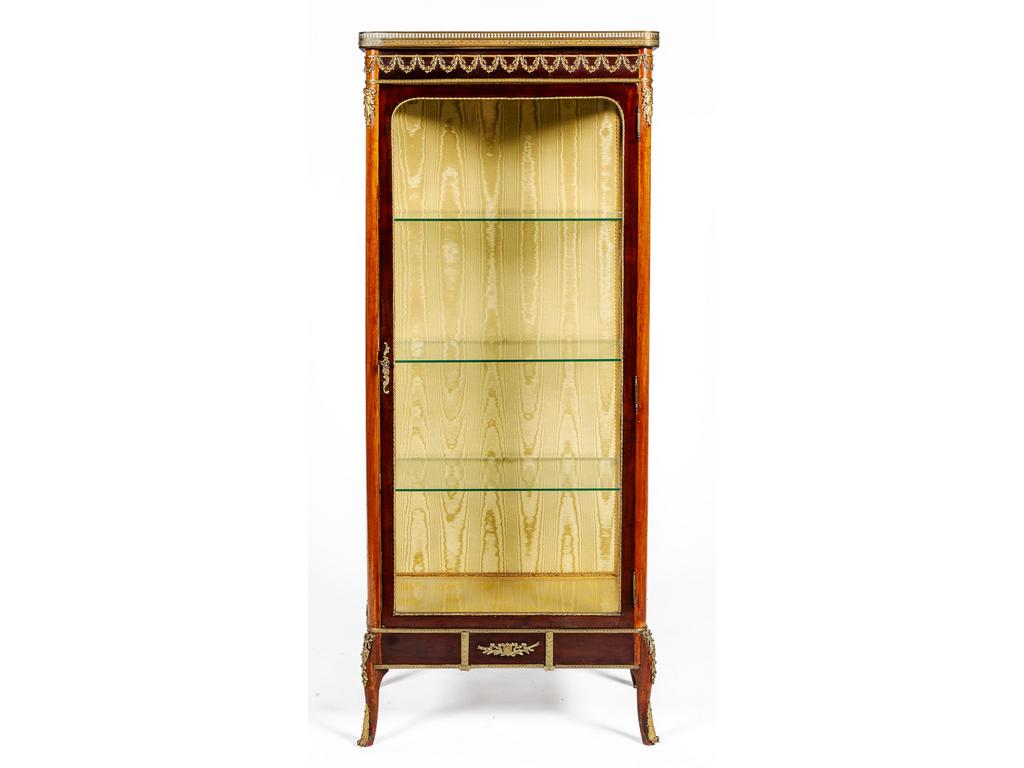 Petite vitrine de style Transition Louis XV - Louis XVI en acajou ornée de guirlande de fleurs et trophée en bronze et laiton. Elle ouvre par une porte et repose sur quatre petits pieds cambrés. Plateau de marbre à galerie. Dim : 146.5 x 61 x 35 cm.