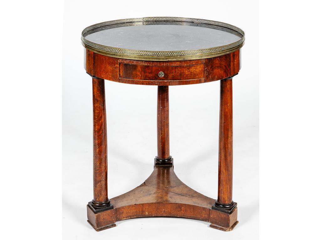 Guéridon circulaire en acajou et placage d'acajou de style Empire ouvrant par un tiroir en ceinture et reposant sur trois pieds colonnes, réunis par une plinthe. Plateau de marbre à galerie de laiton. (Manques). Hauteur : 72 cm. Diamètre : 63.5 cm.