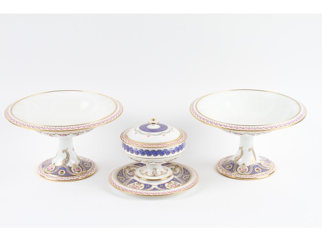 SEVRES : Drageoir et paire de coupes sur pieds en porcelaine blanche à décor peint de fleurs, rubans, en partie sur fond bleu lavande. Le tout réhaussé de dorure. Signés et datés respectivement (18)59 et (18)55. Hauteur des coupes : 16 cm, diamètres : 27 cm. (un pied recollé sans accidents)