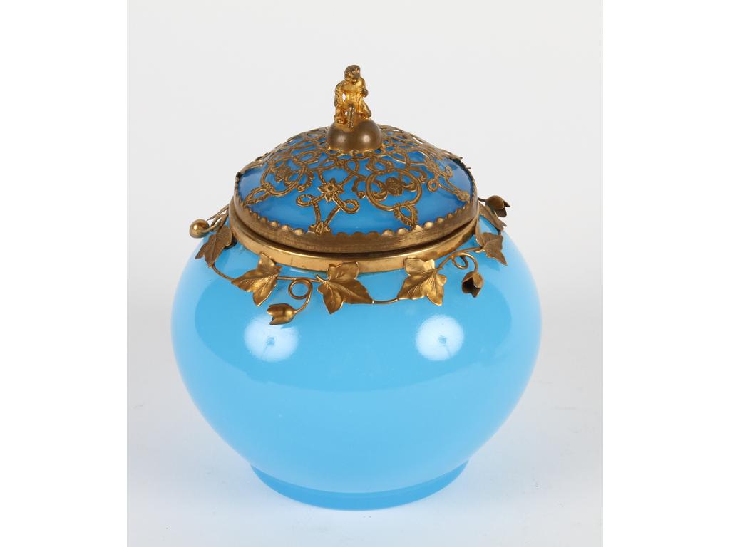 Boite ronde en opaline bleue, le rebord et le couvercle ornés d'une monture en laiton et tôle dorée à décor de lierre, lambrequins et personnage. Bon état. Hauteur : 14 cm.