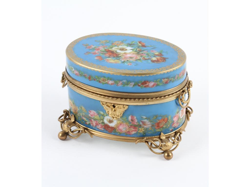 Coffret de forme ovale en porcelaine bleue et à décor peint de bouquets et guirlandes de fleurs. Monture en laiton et tôle dorée. (Bon état général, sans la clé. Eclats à l'intérieur du couvercle). Hauteur : 11 cm.