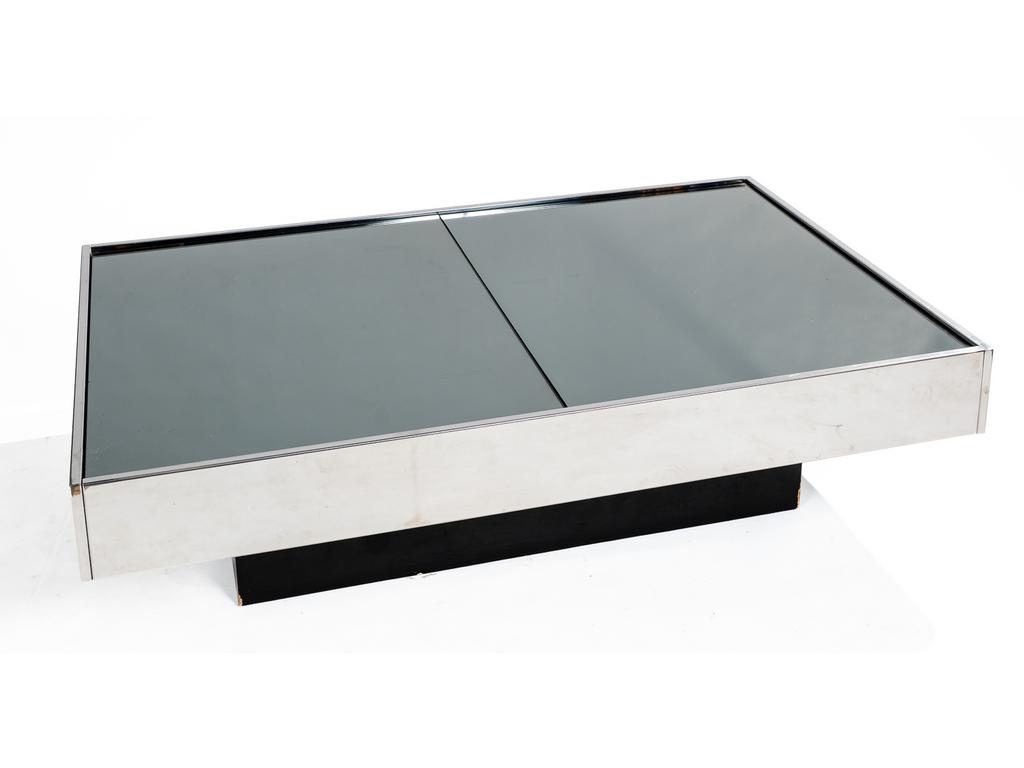 Willy RIZZO (1922-2013) : Table basse rectangulaire reposant sur un caisson en bois stratifié. Les côtés plaqués en métal, le plateau de deux dalles de miroirs teintés noirs. Il s'ouvre au centre par des coulisses et découvre un intérieur (bar) plaqué de miroirs (celui du fond cassé). Hauteur : 35 cm. Dim du plateau : 130 x 80 cm.