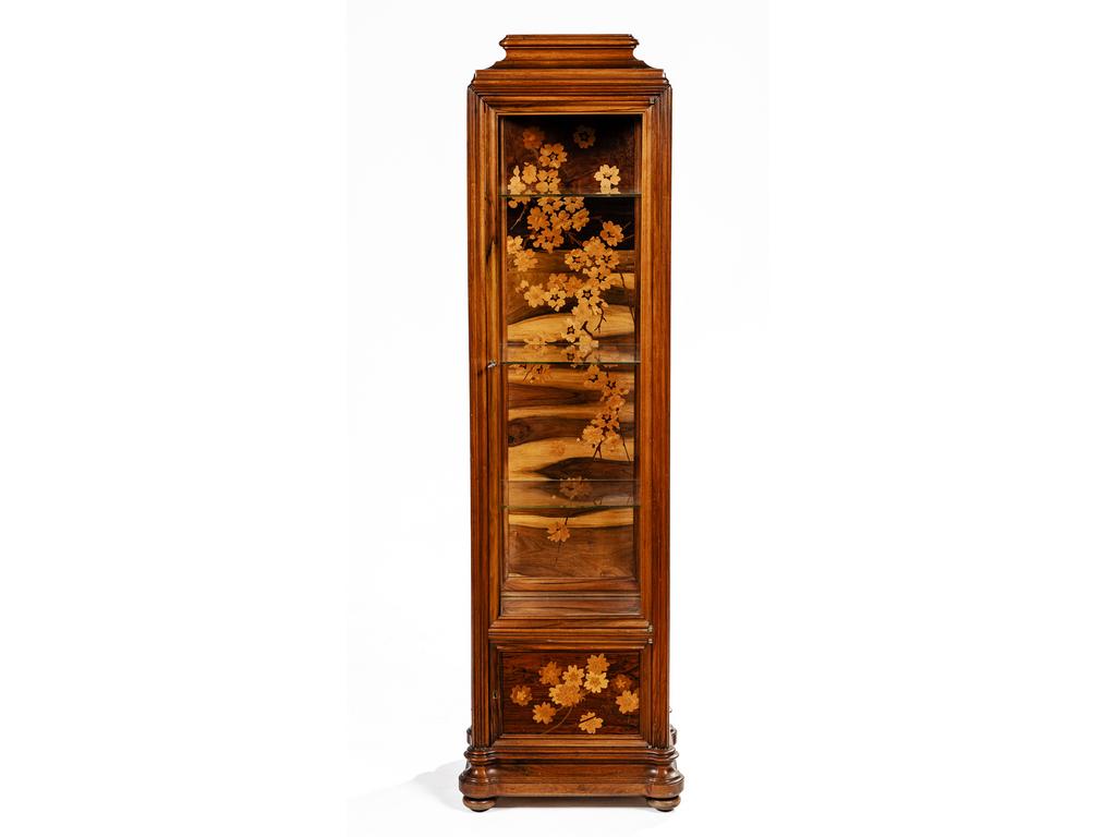 Emile GALLE (1846-1904) :« Branches fleuries de pommier du Japon » daprès le modèle, avec variante de décor sculpté et marqueté, créé initialement vers 1903. Vitrine éclairante de type Colonne en noyer nervuré, mouluré et sculpté. Le corps vertical du meuble souvre par une porte pleine en partie basse surmontée dune grande porte vitrée. La porte pleine, les parties basses latérales et le fond de la vitrine au décor titre réalisé en marqueterie de bois précieux et indigènes. Les parties hautes latérales vitrées , lintérieur équipé de tablettes en verre clair. Les montants, à lévocation de colonne, repose sur une base profondément moulurée reçue par des pieds en sphères aplaties dites aussi miches. Le sommet en doucine et à un gradin central. Probable édition des Etablissements Gallé (1904-1936) dépoque Art Nouveau.