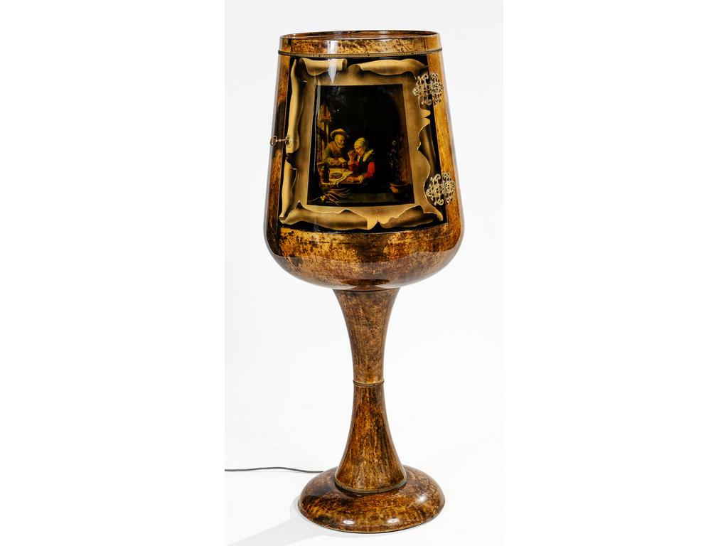 (Attribué à) Aldo TURA (1909-1963), Italie, Années 1950 : Curieux meuble bar en forme de verre à pied en bois et parchemin laqué et verni. Il ouvre par une porte sur charnières ornée d'une scène reproduisant un tableau flamand du XVIIème siècle. Intérieur avec éclairage, plaqué de multiples miroirs avec une étagère en arc de cercle. Hauteur : 127 cm.