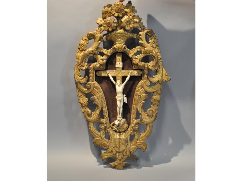 Christ en croix en ivoire sculpté, à ses pieds un memento mori.  Dans un riche encadrement en bois sculpté, ajouré et doré de feuillages, et corbeille de fleurs.  Provence.  XVIIIe siècle.  Hauteur totale : 78 cm.  Hauteur Christ : 19 cm. (petite restauration en partie basse)