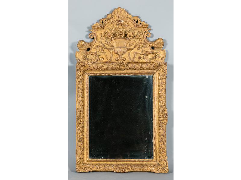 Miroir dans un encadrement en bois doré à décor de rinceaux.  Fronton à décor sculpté d'une corbeille fleurie, palmettes et dauphins.  Milieu XVIIIe siècle.  Dim.: 120x63 cm. (accidents en bas à droite et restaurations aux angles)