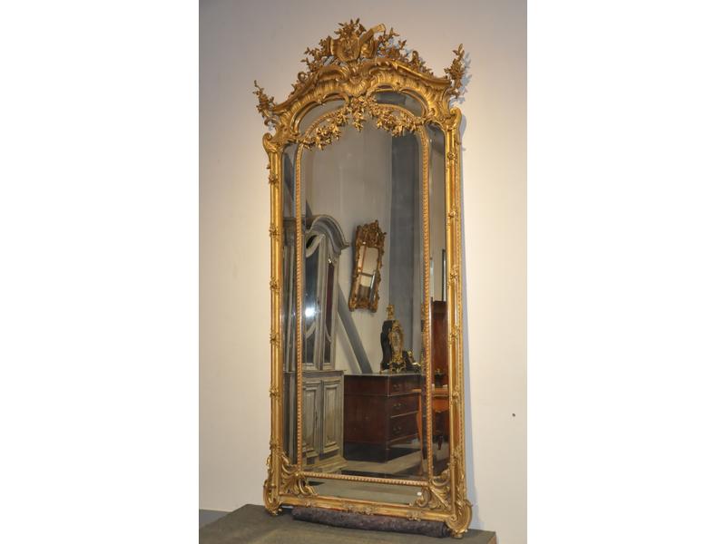 Miroir biseauté dans un encadrement à parecloses en bois et stuc doré à riche décor de style rocaille.  Fronton orné d'attributs musicaux et guirlandes de fleurs.  Dim.: 270x122 cm.