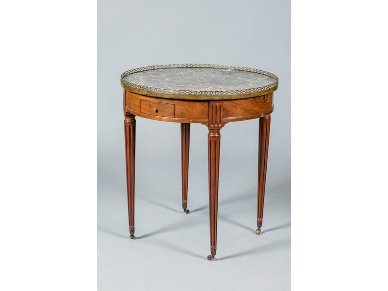 Table bouillote en acajou et placage d'acajou reposant sur quatre pieds cannelés et ouvrant par deux tiroirs et deux tirettes en ceinture.  Plateau de marbre gris entouré d'une galerie ajourée en laiton.  XIXe siècle.  XIXe siècle.  Hauteur : 71 cm.  Diam.: 65 cm.
