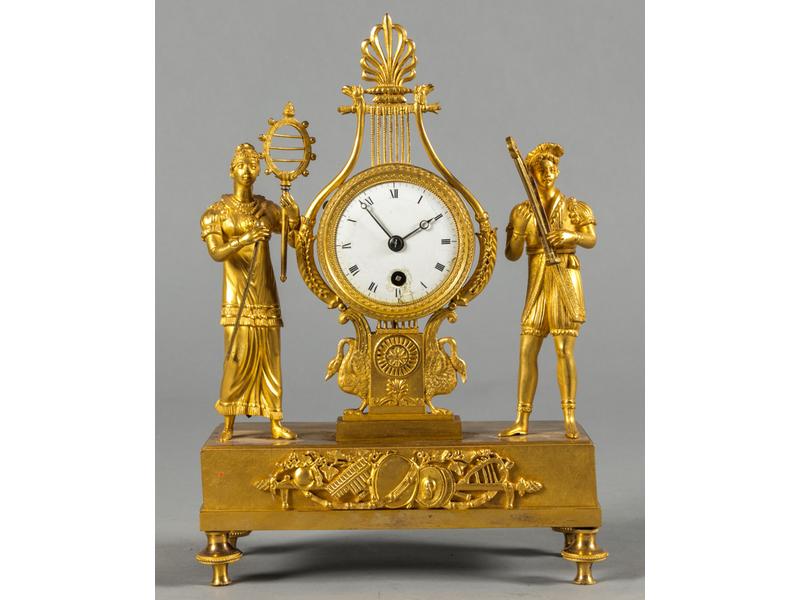 Petite pendule d'époque Directoire en bronze doré ornée d'un joueur de lyre et d'une élégante de part et d'autre d'un cadran émaillé blanc à chiffres romains.  Socle rectangulaire orné d'un trophée d'instruments de musique.  Début XIXe siècle.  Dim. : 29.5x20x8.5 cm.