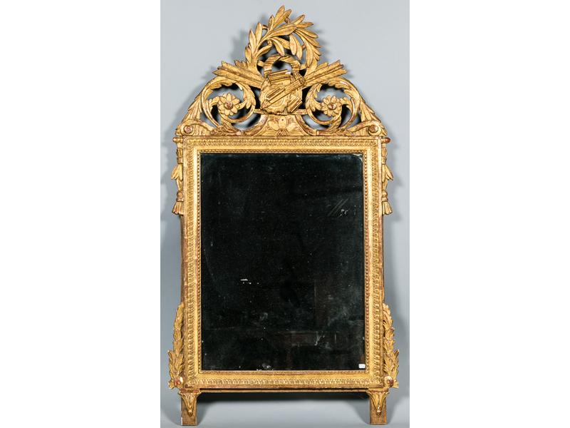 Miroir de forme rectangulaire de style Louis XVI dans un encadrement en bois sculpté et doré surmonté d'un fronton orné d'une couronne de laurier, d'un trophée et de rinceaux feuillagés (quelques sauts de dorure). Dim: 135x75 cm.