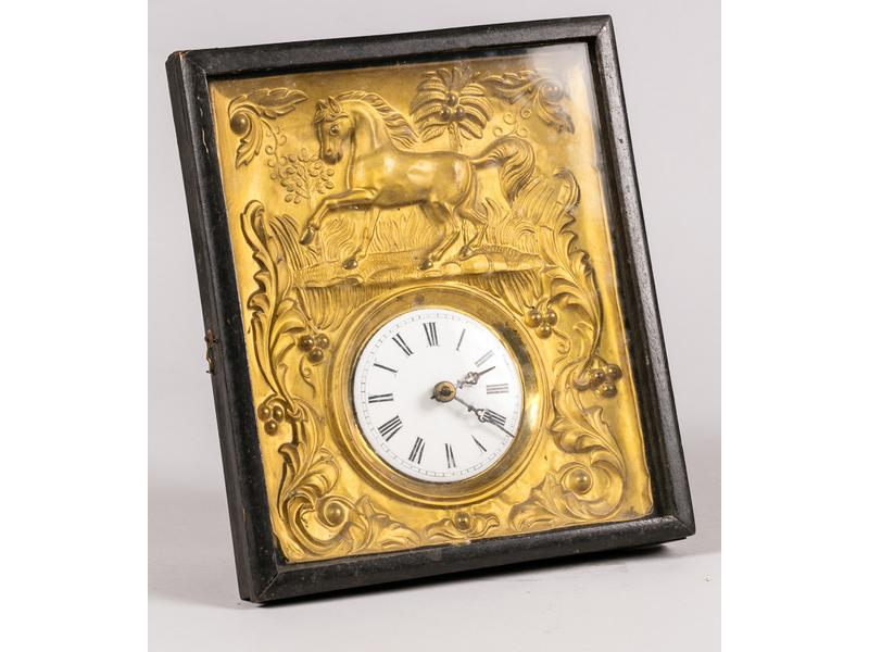 Pendule d'applique de forme rectangulaire présentant un cadran émaillé à chiffres romains dans un entourage en laiton repoussé et doré à décor d'un cheval, palmier et rinceaux feuillagés. Fin XIXe siècle. Dim.: 29x25 cm.