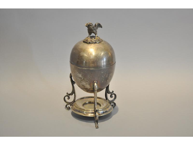 Oeufrier en métal argenté en deux parties reposant sur une base tripode, décoré d'une poule à l'amortissement.  Fin XIXe siècle.  Hauteur : 24 cm.
