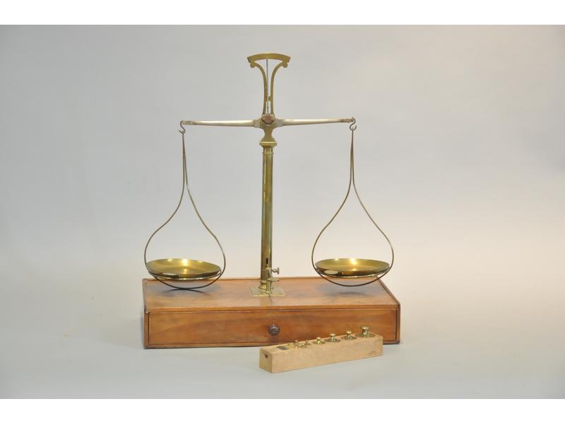 Trébuchet composé d'un socle en noyer ouvrant par un tiroir et de deux plateaux en laiton. On y joint une série complète de poids. Epoque début XXème siècle. Hauteur : 50 cm, longueur : 43 cm