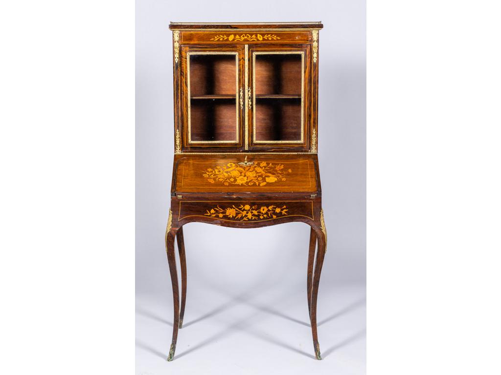 Petit bureau bonheur du jour en placage de palissandre et à décor marqueté de bouquet de fleur. Partie supérieure ouvrant par deux portes vitrées et surmontée d'une galerie en laiton. Il ouvre par un abattant formant pupitre et repose sur quatre petits pieds cambrés. Garniture de bronze. Epoque fin XIX° siècle (petits accidents et manques au placage). Dim: 161x66x42cm.