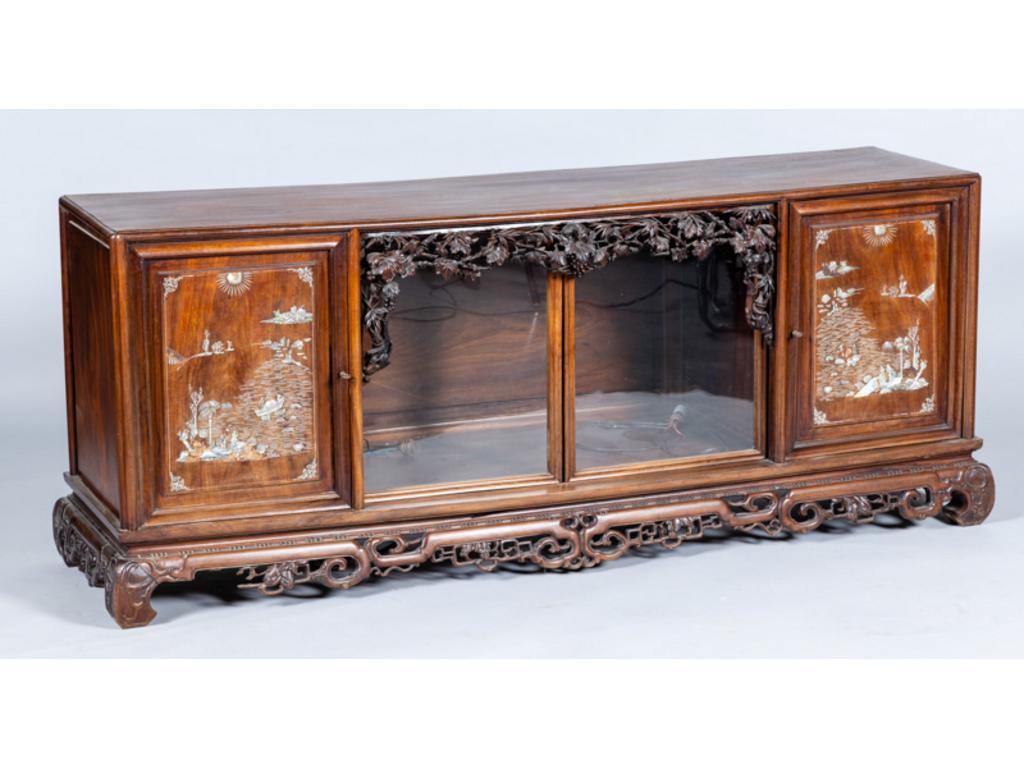 Vitrine basse en bois précieux reposant sur un piètement ajouré à motifs géométriques. Elle ouvre par deux vantaux à décor de chinois dans des paysages et deux portes vitrées encadrées de feuillages et pampres de vigne. Travail Extrême Orient. Fin XIX° siècle. Dim: 69x170x44cm.