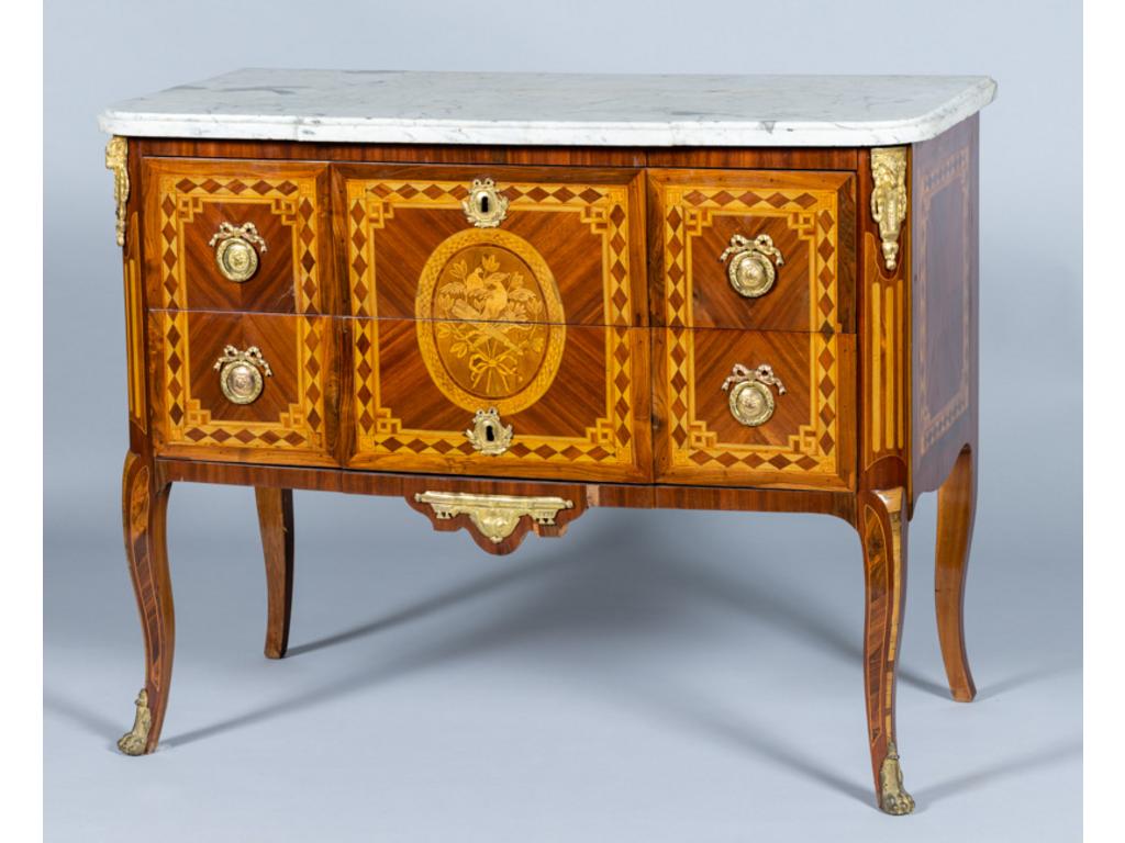 Commode à ressaut de style transition Louis XV Louis XVI en bois de placage à décor marqueté d'un trophée dans un médaillon. Elle ouvre part deux tiroirs sans traverse en façade, montants arrondis à cannelures simulées reposant sur un piètement cambré. Ornementation de bronze. Plateau de marbre blanc. Epoque XIX°. restaurations postérieures. Dim: 89x112x56cm.