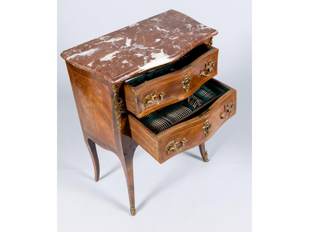 Petite commode de style Louis XV en bois de placage toutes faces galbées ouvrant par deux tiroirs et reposant sur un piètement galbé. Plateau de marbre rouge.. Dim: 83x65x37cm. Intérieur aménagé contenant une ménagère en métal argenté de style Louis XV comprenant 12 grands couverts, 12 cuillères à dessert, 11 fourchettes à dessert, 12 couverts à poisson, 12 fourchettes à huitres, 12 fourchettes à escargot, 12 grands couteaux, 12 couteaux