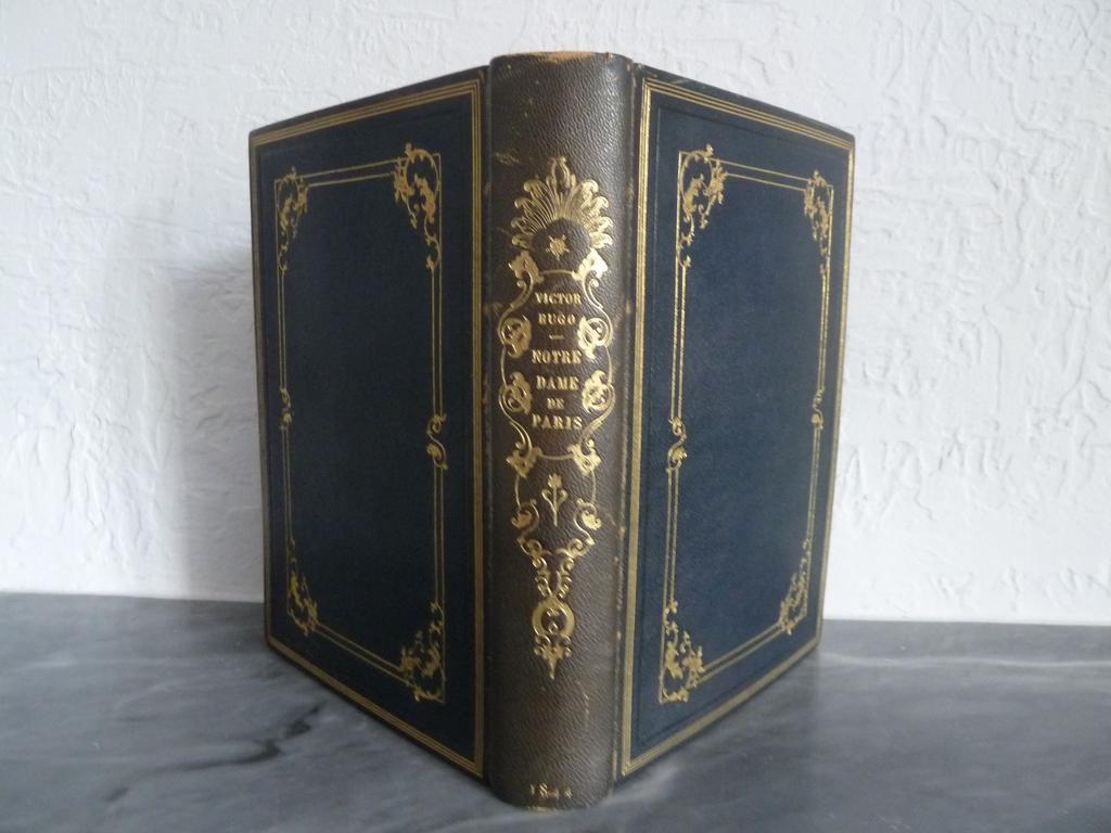 HUGO, Victor : Notre-Dame de Paris. Paris : Perrotin, 1844. Un volume grand in-8°. Reliure plein chagrin bleu nuit, dos lisse orné en long dune composition dorée de filets et rinceaux, double encadrement des plats avec rinceaux dorés aux écoinçons de lencadré intérieure, filet doré sur coupes, roulettes dorées sur chasse, doublure et gardes de papier dominoté, tête dorée. Reliure signée par Marius MAGNIN. Edition illustrée de 55 planches hors-texte dont 21 sur acier et 34 sur bois par MM. E. de Beaumont, L. Boulanger, Daubigny, Tony Johannot, de Lemud, Meissonnier, C. Roqueplan, de Rudder et Steinheil., et de nombreuses figures sur bois in-texte. Au-delà de toutes les spécificités des tirages relevées par les bibliographes, notre exemplaire porte le filet anglais sur la page de titre et le nom des imprimeurs sur les gravures hors-texte sur bois est en petits caractères. (Carteret : III, 299. Vicaire : IV, 260. Brivois : 196). 2 ou 3 feuillets parfaitement restaurées en marge, quasi exempt de rousseurs, dos un brin plus clair, quelques frottis. BON EXEMPLAIRE, de cette « remarquable publication, rare en belle condition, qui peut être recherchée à juste titre comme le prototype du grand texte illustré par les artistes de son temps fameux illustré » (Carteret).